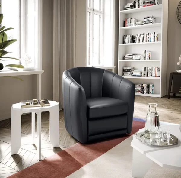 A modern living room with a black armchair, a white side table with books and decor, and a white shelving unit filled with books and items. The room has natural light from two windows.