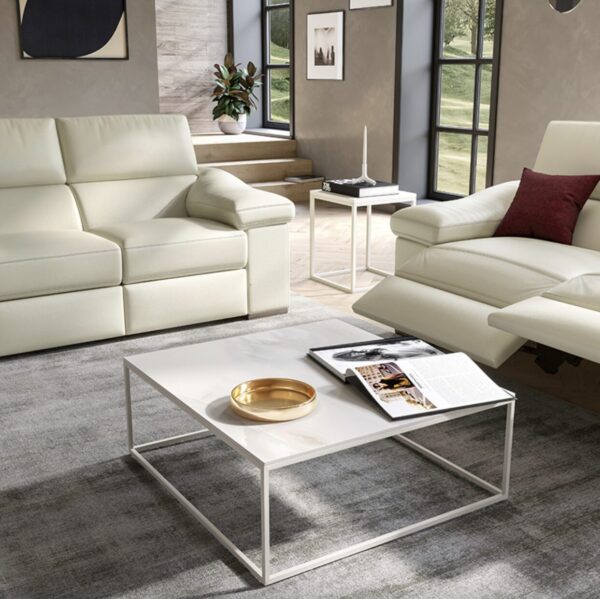 A modern living room with white sofas, a square white coffee table with a gold tray and a magazine, and large windows with outside view.