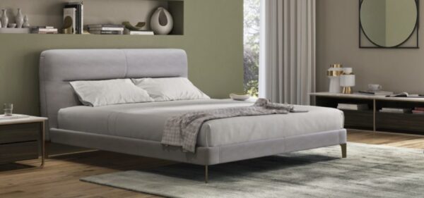 A modern bedroom features a neatly-made LE05 CUT light gray bed, flanked by nightstands with books and a lamp. The room also includes a rug, a wall mirror, and a window with curtains allowing natural light in.