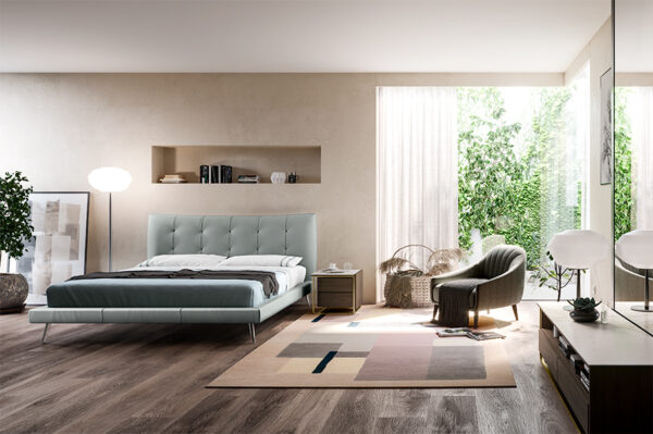 A modern bedroom with a large LE03 GALATTICO, cushioned headboard, gray armchair, and colorful rug. Natural light enters through a large window, and a potted plant is on the left.
