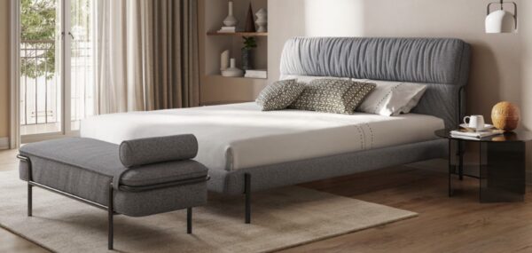A modern bedroom with a gray upholstered bed, white bedding, and patterned pillows. A matching gray bench is at the foot of the bed. The room features a black side table, light wood floors, and beige walls.